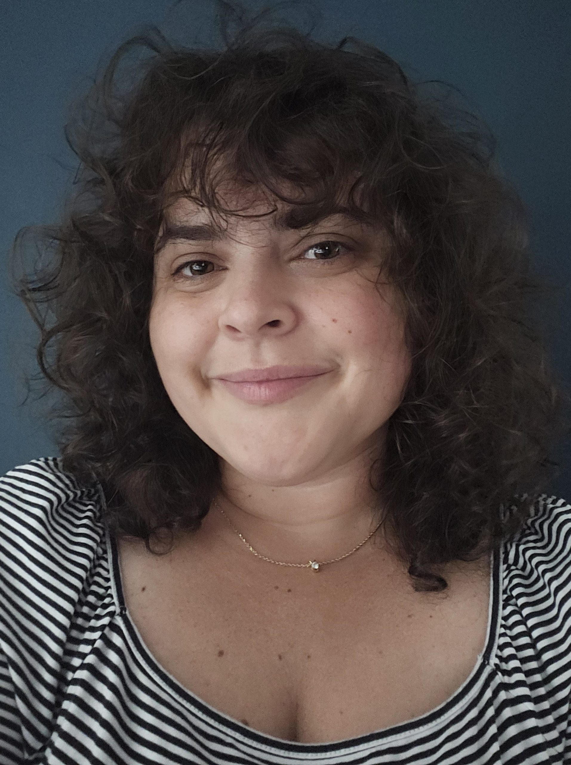 A photograph of the author, a white woman with dark brown curly hair, brown eyes, and freckled skin. She is wearing a gold necklace, a black and white striped shirt, and is standing in front of a green background.